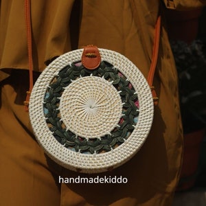 Puede incluir: Bolso bandolera redondo tejido en tonos blancos y naturales. El bolso presenta un dise&ntilde;o central tejido, rodeado por un dise&ntilde;o floral y verde oscuro. Tiene una correa y cierre de cuero marr&oacute;n. El texto "handmadekiddo" es visible.