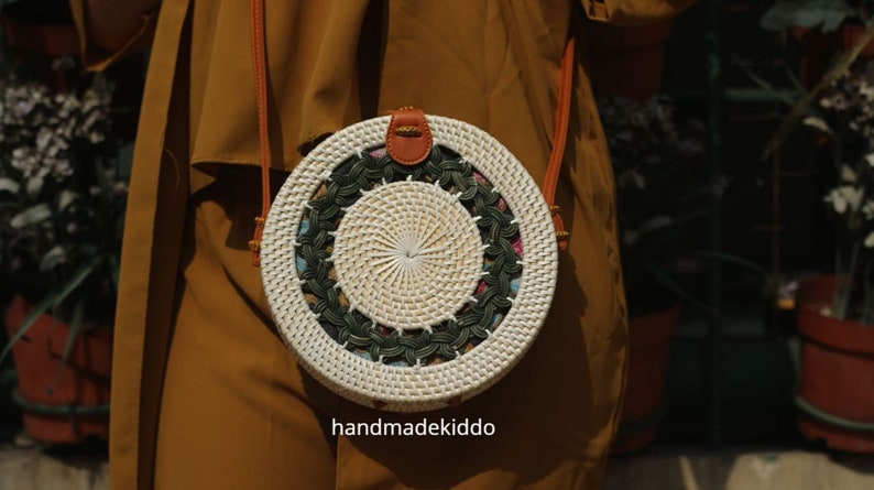 Puede incluir: Bolso bandolera redondo tejido en tonos blancos y naturales. El bolso presenta un dise&ntilde;o central tejido, rodeado por un dise&ntilde;o floral y verde oscuro. Tiene una correa y cierre de cuero marr&oacute;n. El texto "handmadekiddo" es visible.