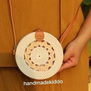 May include: A round, white and beige woven bag with a brown leather strap. The bag has a circular design with a smaller circle in the center. The bag is handmade and has the text "handmadekiddo" printed on the bottom.