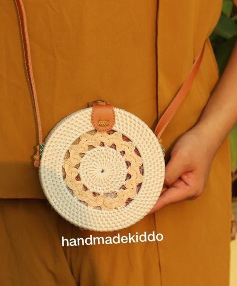 May include: A round, white and beige woven bag with a brown leather strap. The bag has a circular design with a smaller circle in the center. The bag is handmade and has the text "handmadekiddo" printed on the bottom.