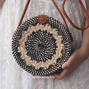 May include: A round, woven bag with a black and white pattern. The bag has a brown leather strap and is being held by a person.