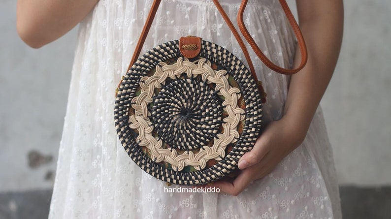 May include: A round, woven bag with a black and white pattern. The bag has a brown leather strap and is being held by a person.