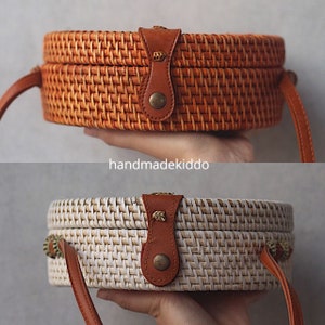 May include: Two round woven rattan bags with brown leather straps and gold accents. The top bag is brown and the bottom bag is white.