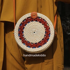 Puede incluir: Un bolso redondo de rat&aacute;n tejido, con un centro color crema y un patr&oacute;n decorativo en rojo y azul. El bolso tiene una correa de cuero marr&oacute;n y un peque&ntilde;o detalle de cuero en la parte superior. El texto "handmadekiddo" es visible en la parte inferior.