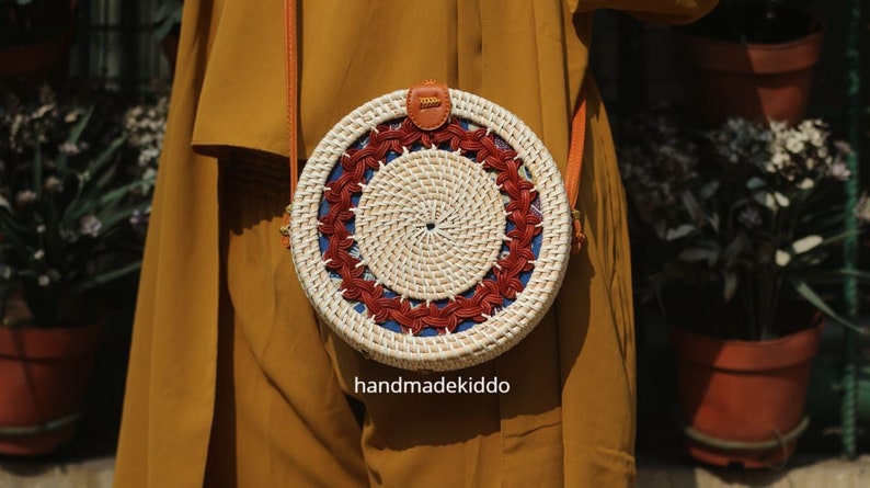 Puede incluir: Un bolso redondo de rat&aacute;n tejido, con un centro color crema y un patr&oacute;n decorativo en rojo y azul. El bolso tiene una correa de cuero marr&oacute;n y un peque&ntilde;o detalle de cuero en la parte superior. El texto "handmadekiddo" es visible en la parte inferior.
