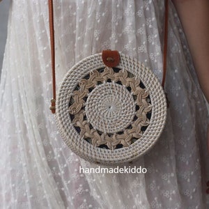 May include: White round woven rattan bag with a brown leather strap. The bag has a circular pattern with a woven design.