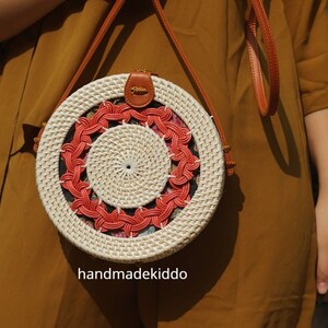 Puede incluir: Bolso redondo de rat&aacute;n tejido con un dise&ntilde;o en espiral. El bolso presenta una base de color crema con un patr&oacute;n en espiral rojo y crema. Tiene una correa de cuero marr&oacute;n y un peque&ntilde;o cierre de cuero. El texto "handmadekiddo" es visible.