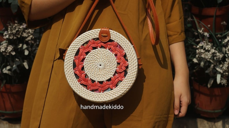 Puede incluir: Bolso redondo de rat&aacute;n tejido con un dise&ntilde;o en espiral. El bolso presenta una base de color crema con un patr&oacute;n en espiral rojo y crema. Tiene una correa de cuero marr&oacute;n y un peque&ntilde;o cierre de cuero. El texto "handmadekiddo" es visible.
