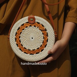 Puede incluir: Bolso bandolera redondo de rat&aacute;n tejido, con centro blanco y borde naranja decorativo. Cuenta con una correa de cuero marr&oacute;n y un peque&ntilde;o detalle de cuero en la parte superior. El texto "handmadekiddo" es visible.