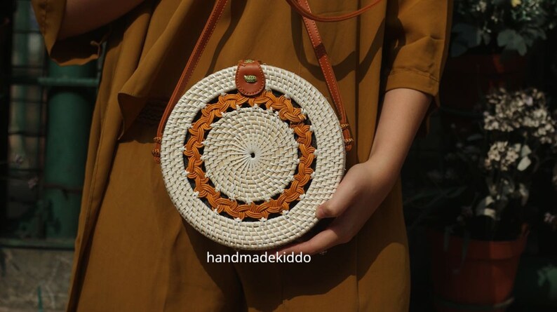 Puede incluir: Bolso bandolera redondo de rat&aacute;n tejido, con centro blanco y borde naranja decorativo. Cuenta con una correa de cuero marr&oacute;n y un peque&ntilde;o detalle de cuero en la parte superior. El texto "handmadekiddo" es visible.