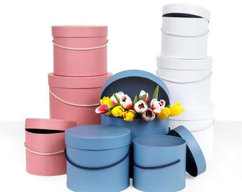May include: A collection of round cardboard gift boxes in pink, blue, and white, featuring matching rope handles. One blue box is open and filled with a bouquet of yellow, white, and pink tulips. The boxes are arranged in various sizes.