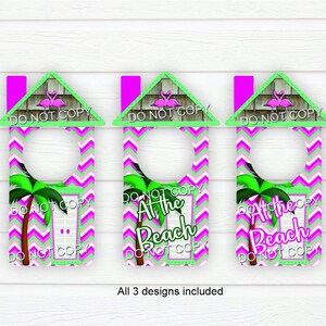 May include: Three pink and green door hangers with a house shape and a flamingo design. The text on the hangers says "At the Beach".