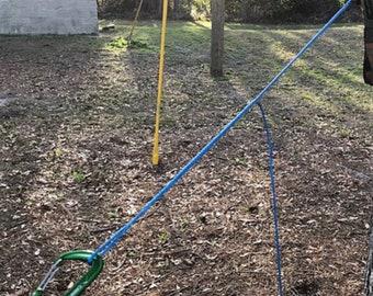 May include: A blue rope is tied to a tree branch and a green carabiner clip is attached to the rope. The rope is also tied to a yellow pole in the background.