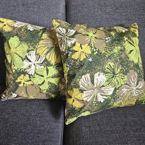 Peut inclure: Deux coussins carrés avec un motif floral. Les coussins présentent un fond vert foncé avec de grandes fleurs stylisées dans des tons de jaune, blanc et vert clair. Ils sont posés sur un canapé gris.