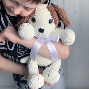 Peut inclure: Un enfant tient un jouet en peluche en crochet en forme de chien avec une fourrure brune et blanche. Le chien a un ruban blanc autour du cou et de grands yeux bruns.