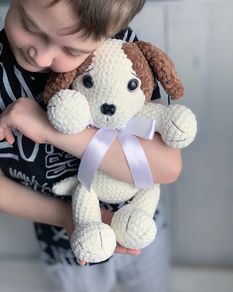 Peut inclure: Un enfant tient un jouet en peluche en crochet en forme de chien avec une fourrure brune et blanche. Le chien a un ruban blanc autour du cou et de grands yeux bruns.