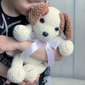 Peut inclure: Un enfant tient un jouet en peluche en crochet blanc et marron en forme de chien avec un ruban blanc autour du cou. Le chien a de grands yeux noirs et un nez brun.
