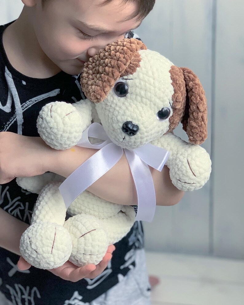 Peut inclure: Un enfant tient un jouet en peluche en crochet blanc et marron en forme de chien avec un ruban blanc autour du cou. Le chien a de grands yeux noirs et un nez brun.