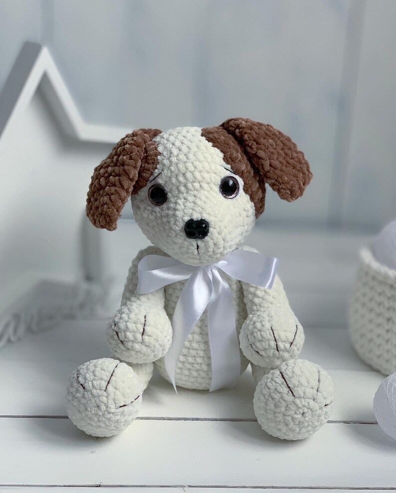 Peut inclure: Un jouet en crochet pour chien blanc avec des oreilles brunes et un ruban de satin blanc. Le chien a des yeux noirs et un nez noir.