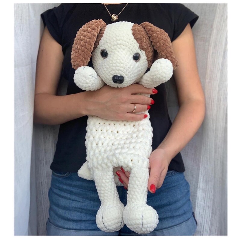Peut inclure: Un jouet en peluche de chien en crochet blanc et marron avec des yeux marrons et un nez noir. Le jouet est tenu par une personne.
