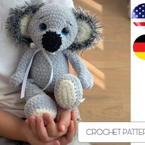 May include: A crocheted koala plush toy with a white ribbon, gray fur, and black eyes. The toy is being held by a person's hands. CROCHET PATTERN is written on a white button below the toy.
