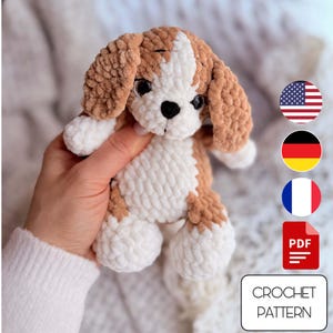 Peut inclure: Un chiot au crochet fait à la main, marron et blanc. Le jouet en peluche a de grandes oreilles tombantes, une bande blanche sur le visage et des yeux noirs. L'image comprend une icône PDF et le texte "CROCHET PATTERN".