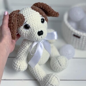 Peut inclure: Un chien en crochet blanc avec des oreilles brunes et un n&oelig;ud en satin blanc. Le chien a des yeux noirs et un nez noir.