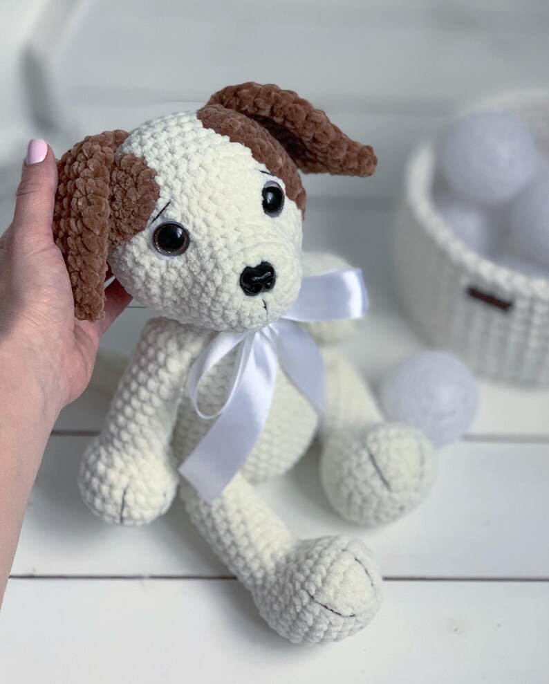 Peut inclure: Un chien en crochet blanc avec des oreilles brunes et un n&oelig;ud en satin blanc. Le chien a des yeux noirs et un nez noir.