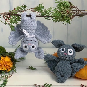 Puede incluir: Dos murciélagos de crochet colgando de una rama. Un murciélago es gris con detalles blancos y el otro es gris oscuro.