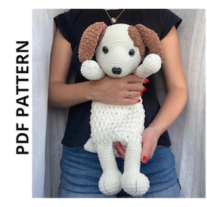 Può includere: Un giocattolo di peluche di cane bianco all'uncinetto con orecchie marroni. Il cane è tenuto da una persona che indossa una maglietta nera e jeans blu.