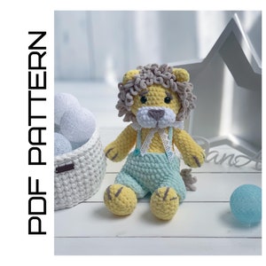 May include: A crocheted lion toy with a yellow body, gray mane, and blue overalls. The lion is sitting on a white surface with a white basket and a blue ball.