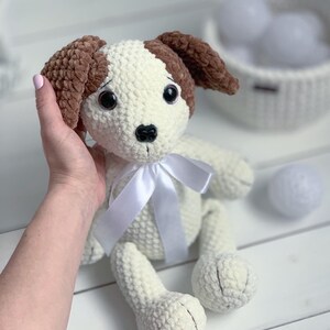 Peut inclure: Jouet en crochet pour chien blanc avec des oreilles brunes et un ruban blanc. Le chien a des yeux noirs et un nez noir.