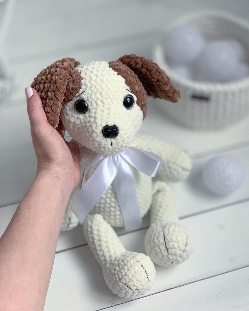 Peut inclure: Jouet en crochet pour chien blanc avec des oreilles brunes et un ruban blanc. Le chien a des yeux noirs et un nez noir.
