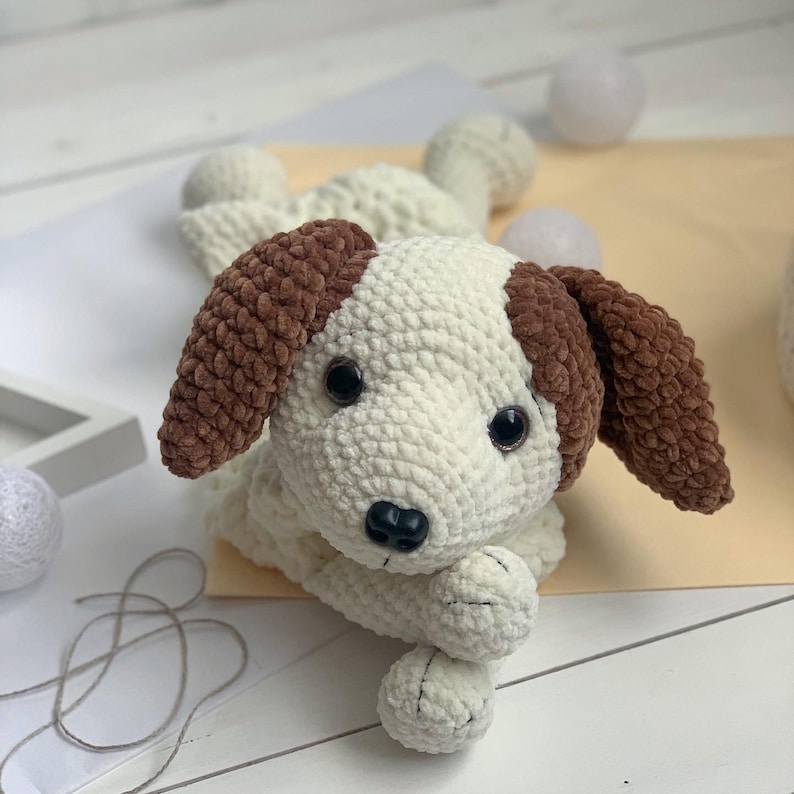Peut inclure: Un chien en peluche crochet&eacute; avec une fourrure blanche et des oreilles brunes. Le chien est couch&eacute; sur le c&ocirc;t&eacute;, la t&ecirc;te reposant sur ses pattes.