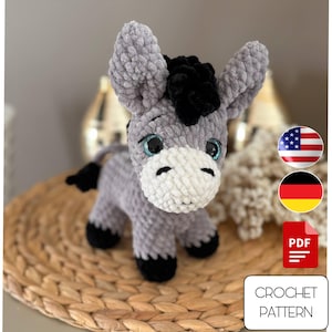 Könnte beinhalten: Ein handgehäkeltes graues Eselchen mit schwarzer Mähne, Schwanz und Hufen sowie einer weißen Schnauze. Der Esel hat große, blaue Augen und sitzt auf einer gewebten Matte. Das Bild enthält ein PDF-Symbol und den Text "CROCHET PATTERN".