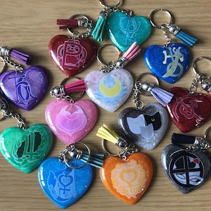 May include: A collection of heart-shaped keychains in various colors, including red, blue, and green, with glittery surfaces. Each keychain features a unique design or symbol, and is adorned with a tassel.
