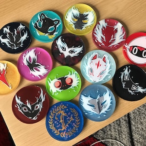 Op de afbeelding: Een verzameling ronde hars coasters met diverse anime-stijl karakterontwerpen. De coasters tonen afbeeldingen van gestileerde dieren en symbolen in zwart, wit, rood, geel, groen, blauw en roze. De coasters zijn gerangschikt op een lichtbruin oppervlak.