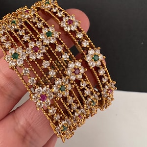 Peut inclure: Un ensemble de bracelets rigides dorés présentant un motif floral répétitif. Chaque fleur est incrustée de pierres blanches transparentes et centrée par une petite pierre ronde rouge ou verte. La surface métallique a une finition perlée.