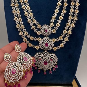 May include: A multi-strand necklace and matching earrings set featuring silver-toned metal, clear pavé stones, and teardrop-shaped pink gemstones. The necklace has three layers with floral motifs and a large central pendant with dangling pink stones.