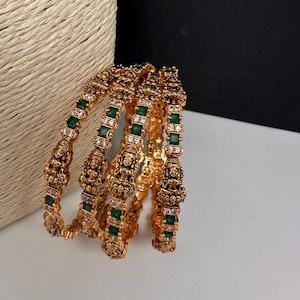 May include: A set of four gold-toned bangles featuring intricate embossed deity figures, alternating with rectangular green stones and small clear white stones. The metalwork displays a textured, antique finish with detailed craftsmanship.