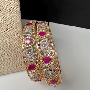 May include: Two gold-toned bangles featuring an intricate openwork design encrusted with small clear stones. Each bangle is accented with evenly spaced, teardrop-shaped fuchsia pink stones set within the metallic framework of the jewelry.