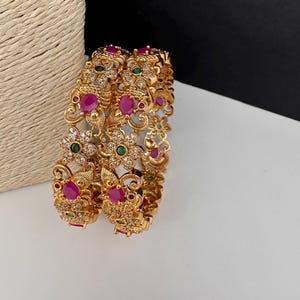 May include: Two gold-tone bangles featuring intricate metalwork, embedded with oval-shaped pink stones, small round green stones, and clear white crystals arranged in floral and peacock-inspired patterns across the surface of the metal bands.
