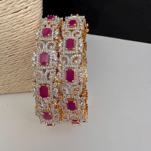 May include: Two gold-tone bangles featuring rectangular, deep pink stones set in a repeating pattern. The metal framework is intricately detailed with numerous small, clear, sparkling stones surrounding each pink stone in a decorative arrangement.