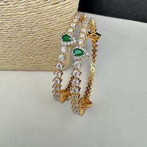 May include: A pair of gold-tone bangles featuring a continuous row of clear round-cut stones and a prominent teardrop-shaped green stone accent set within a cluster of smaller clear stones on each piece.