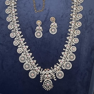 May include: A dazzling necklace and earring set. The necklace and earrings feature a floral design with clear and pink gemstones. The necklace has a gold chain and a pendant with a detailed design. The jewelry is set against a dark blue background.