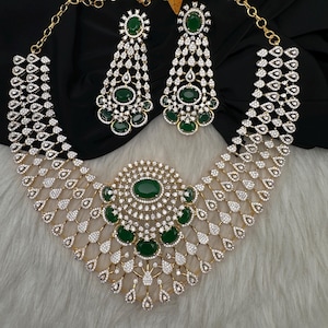 May include: A gold-toned necklace and matching drop earring set featuring oval-cut dark green stones surrounded by clear pavé-set crystals. The necklace has a wide, multi-layered design with a central circular motif and a delicate chain link.