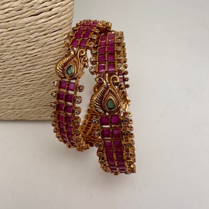 May include: Two gold-toned bangles with a repeating pattern of square, ruby-colored stones. The bangles feature ornate peacock-shaped accents with green and clear stones. The jewelry is displayed on a white surface.