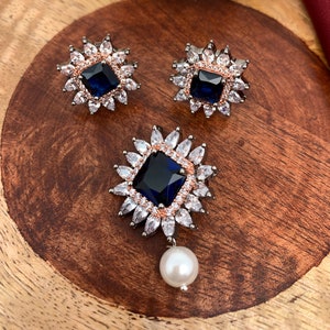 May include: A jewelry set featuring a pendant and matching earrings. The pendant and earrings have a square, dark blue center stone surrounded by white and rose-gold colored accents. The pendant has a dangling white pearl.
