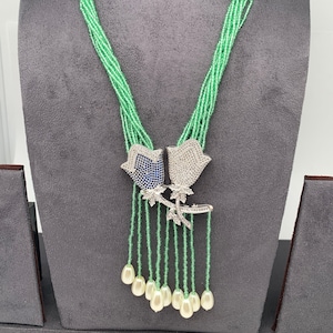 May include: A beaded necklace with a light green strand design. The necklace features two flower pendants with blue and white accents, and dangling white teardrop-shaped beads. The necklace is displayed on a gray surface.