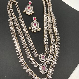 May include: A three-strand necklace and matching earrings set. The jewelry features a gold-tone metal with clear, sparkling stones and ruby-colored center stones. The earrings have a pearl drop design.